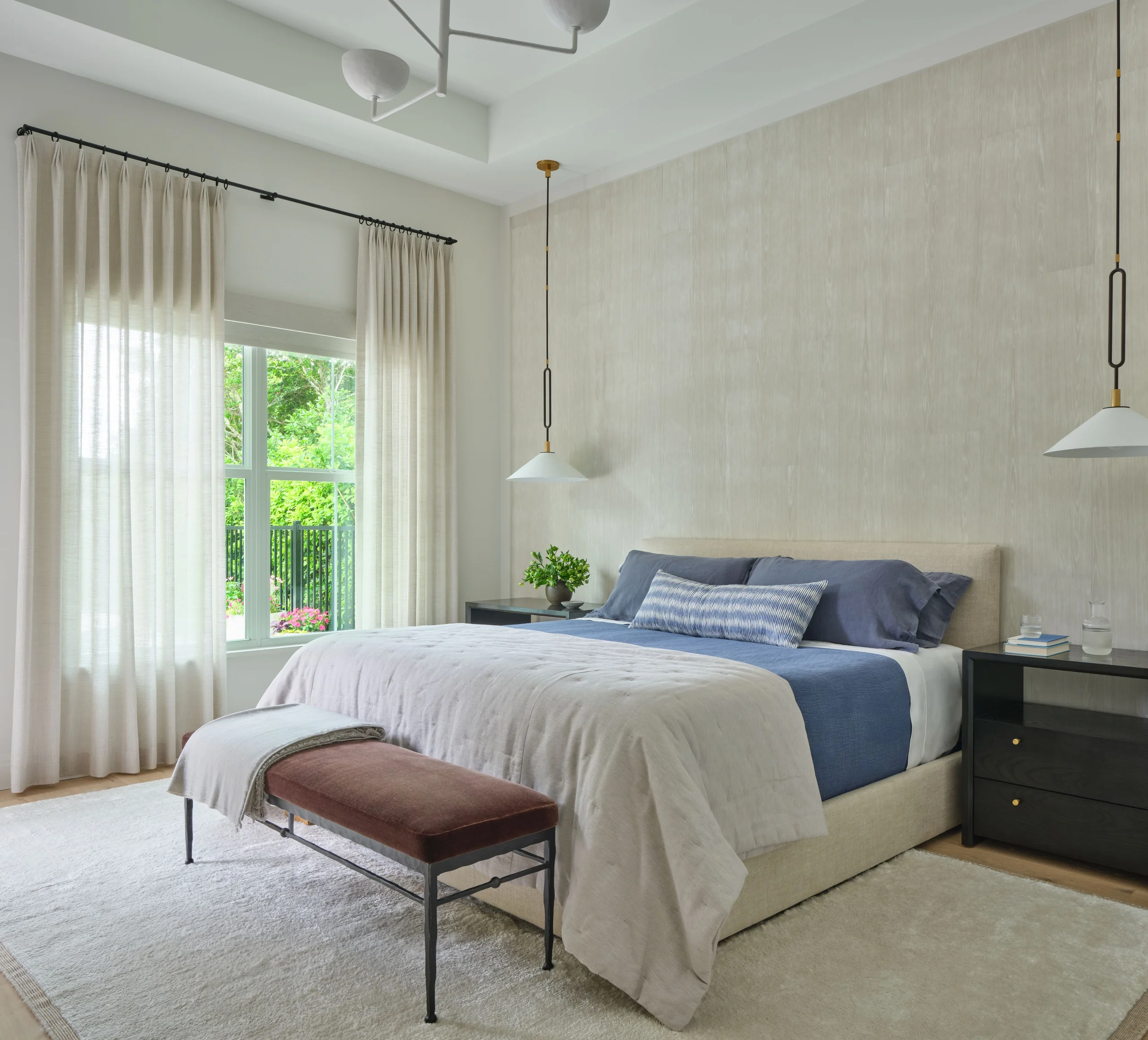 Modern bedroom with a large beige upholstered bed dressed in blue and beige bedding, two dark wood nightstands with books and glassware, hanging pendant lights, a bench with a brown cushion and a beige throw, a large window with sheer white curtains, and a light-colored area rug on a wooden floor.