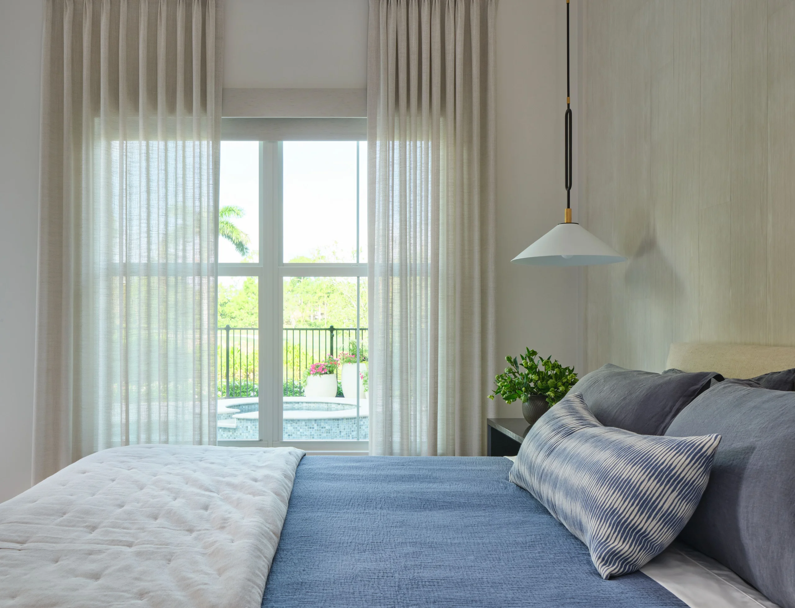 Modern bedroom with a bed dressed in blue bedding and patterned pillow, light beige curtains framing a window showing an outdoor pool and greenery, a small plant on a dark bedside table, and a white pendant lamp hanging above.