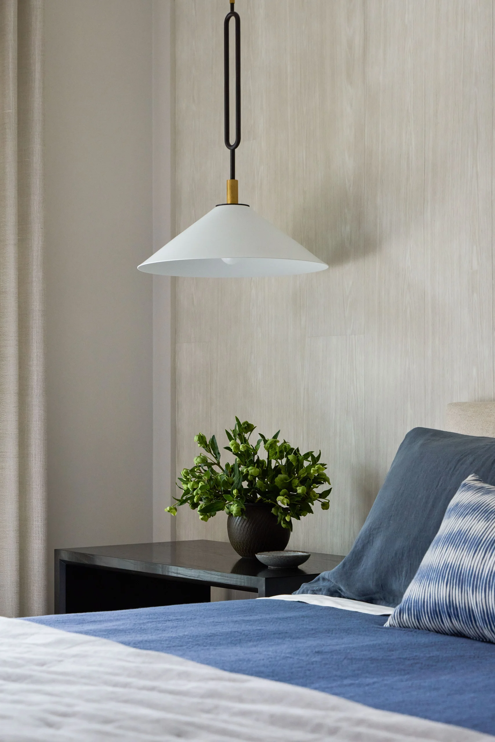 Modern bedroom corner showing part of a bed with blue and white bedding, a black bedside table with a green leafy plant in a dark vase, a small gray dish, and a white pendant light hanging above against a light wooden wall.
