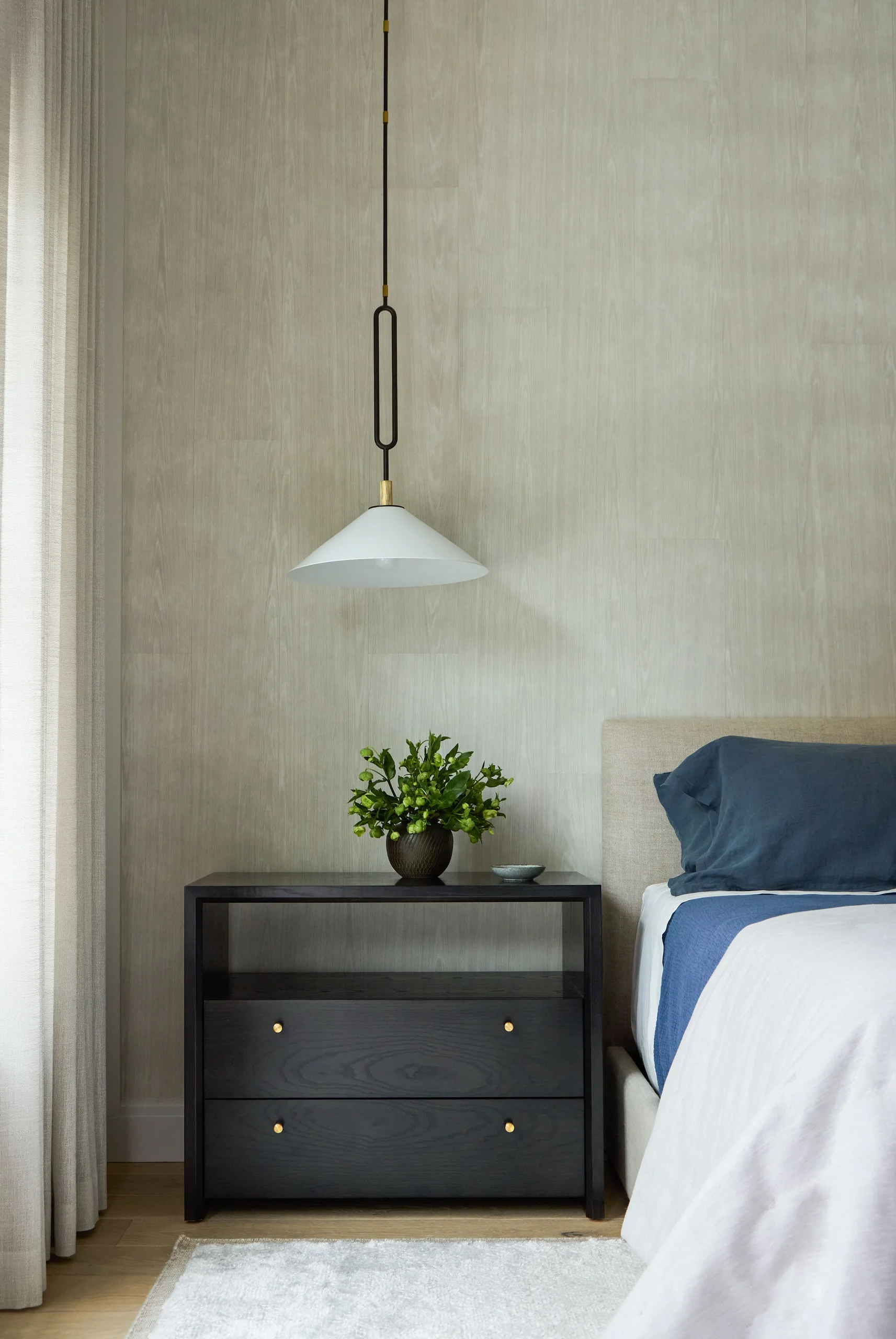 Modern bedroom corner with a dark wood nightstand featuring two drawers with brass knobs, topped with a green leafy plant in a pot and a small bowl. Above it hangs a white cone-shaped pendant light with black and brass details. Part of a beige upholstered bed with blue bedding is visible on the right, and light sheer curtains hang to the left against a textured light gray wall.