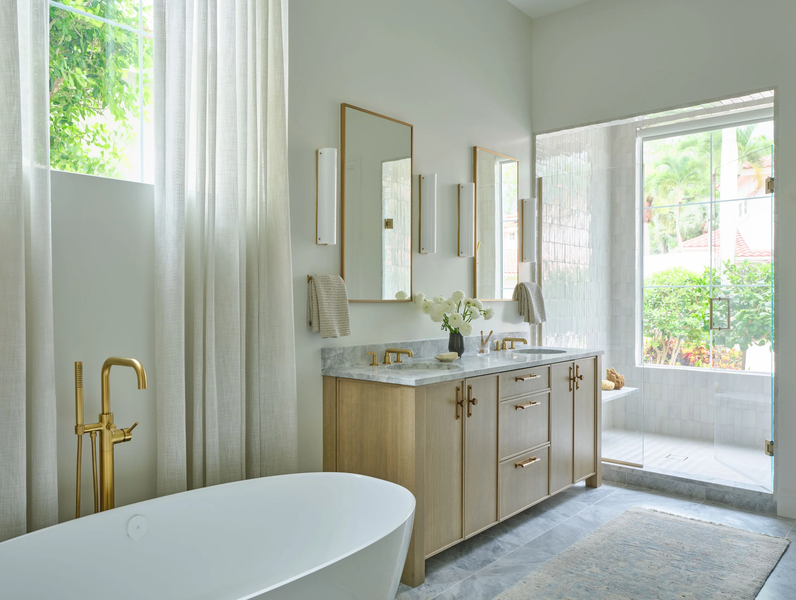 Modern bathroom with a white freestanding bathtub and gold faucet near a large window with sheer white curtains, a wooden vanity with marble countertop featuring two gold faucets, rectangular mirrors, wall-mounted lights, and towels, next to a glass-enclosed shower with a view of green plants outside.