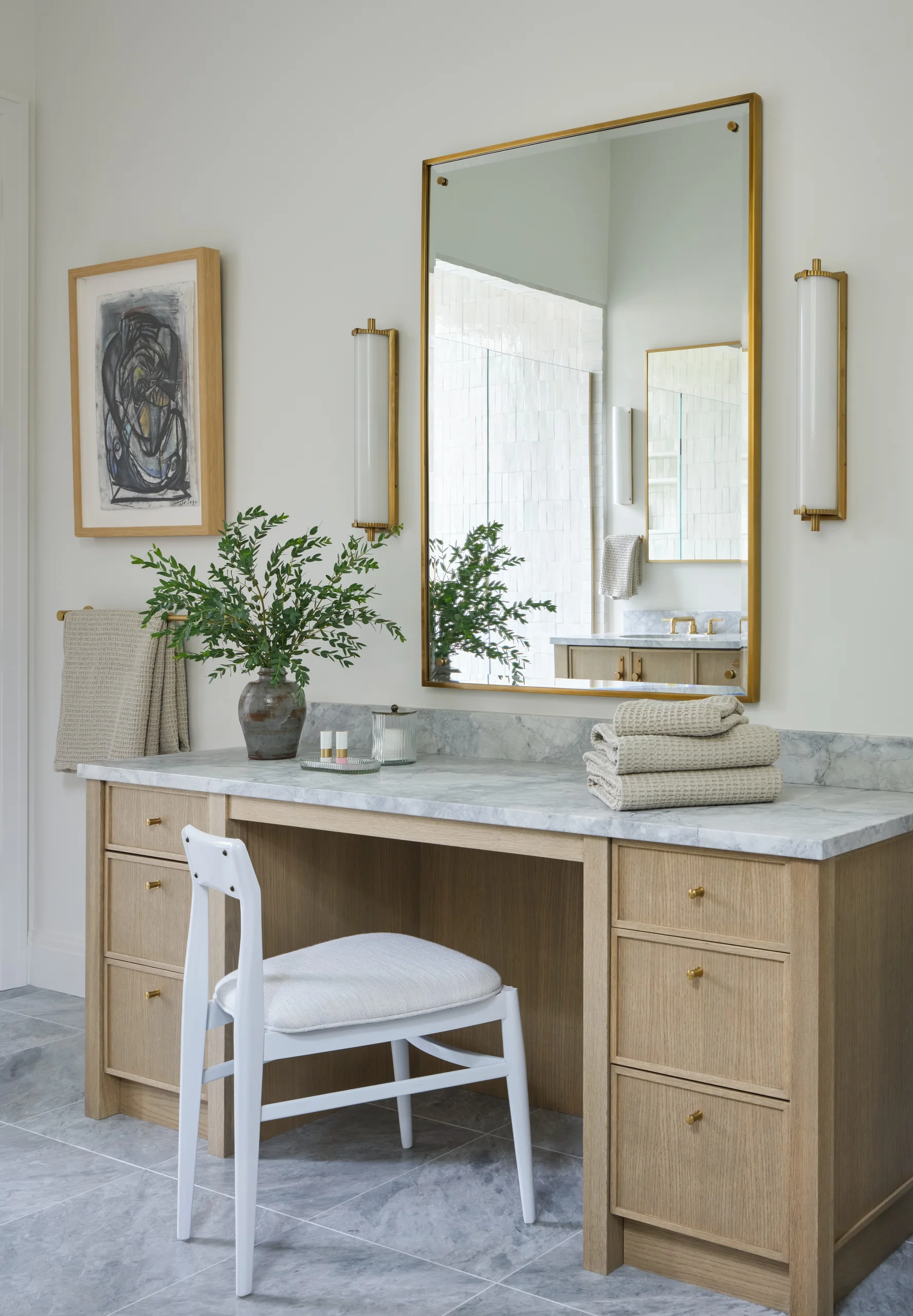 Contemporary bathroom vanity area with a marble countertop, light wood cabinetry, a large rectangular gold-framed mirror, two vertical brass sconces, a white cushioned chair, folded beige towels, a green leafy plant in a ceramic vase, and abstract artwork on the wall.