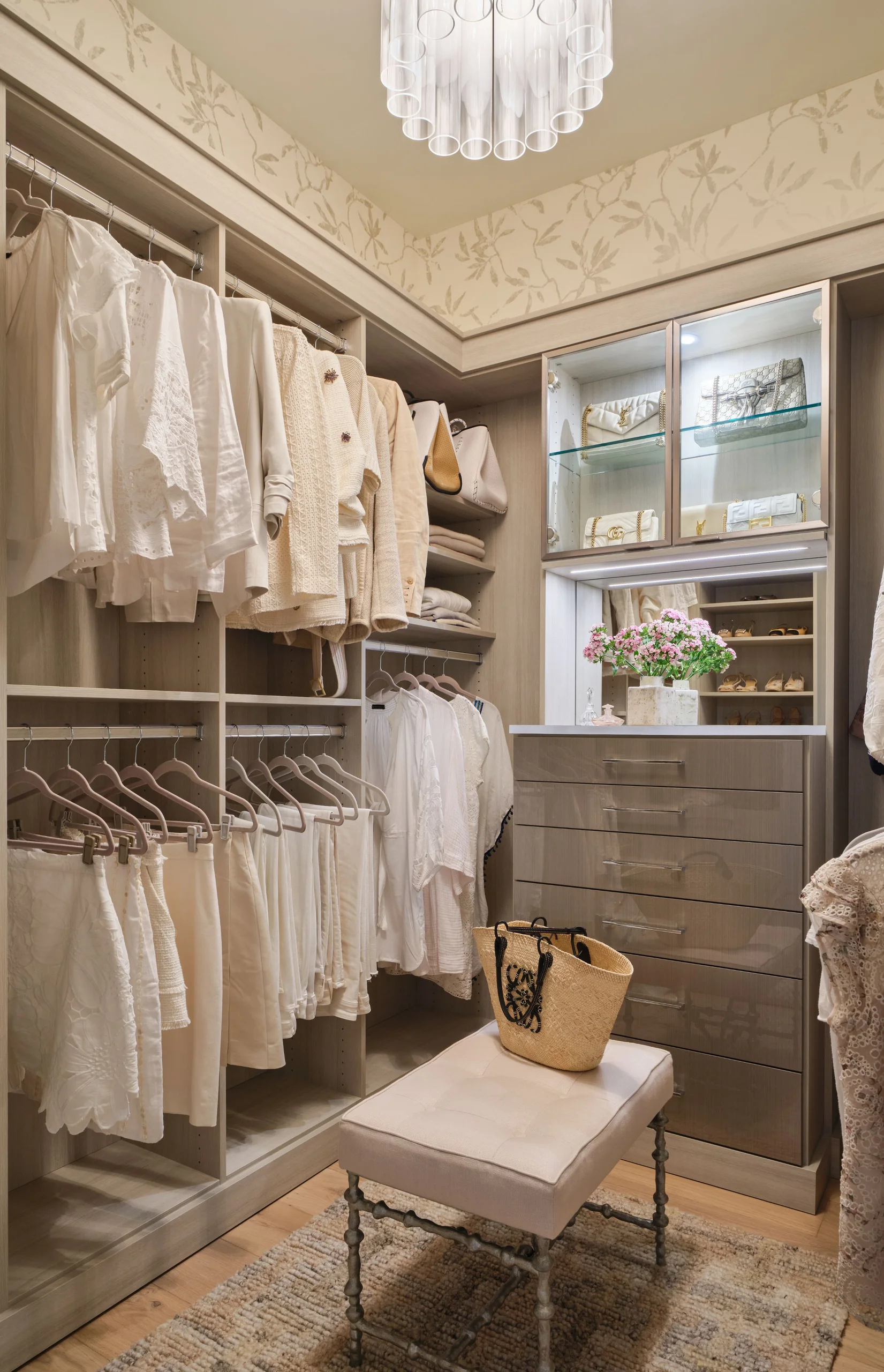 Elegant walk-in closet featuring beige and white clothes neatly hanging on rods, shelves with folded items and handbags, glass-front cabinet displaying luxury purses, a vanity area with a vase of pink flowers, and a cushioned bench with a woven straw bag on a textured rug.