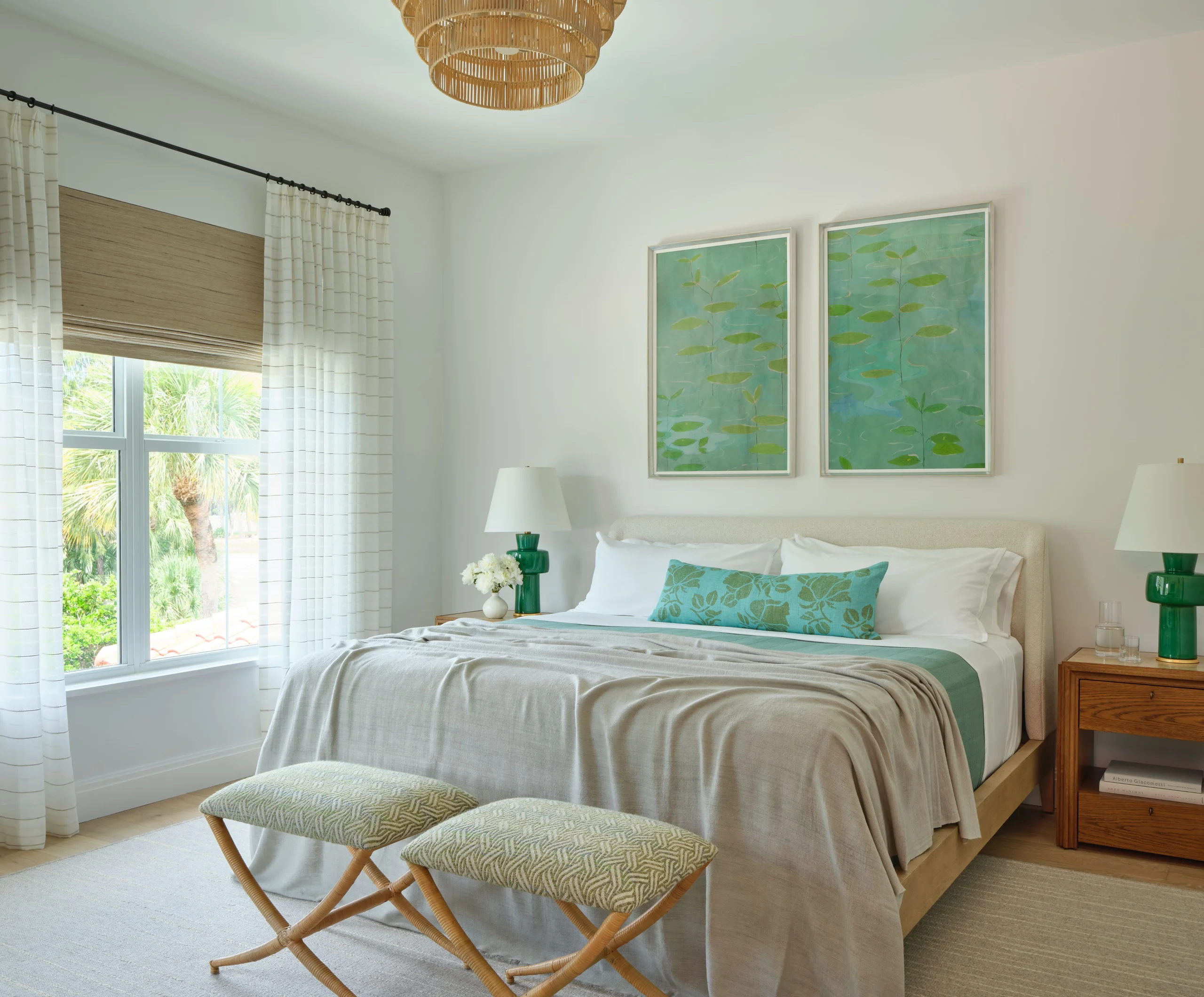 Bright bedroom with a large bed covered in light gray bedding and teal accents, flanked by wooden nightstands with white lamps and floral decorations, two matching patterned stools at the foot of the bed, large window with sheer white curtains and a wooden blind, and two framed green botanical paintings above the headboard.