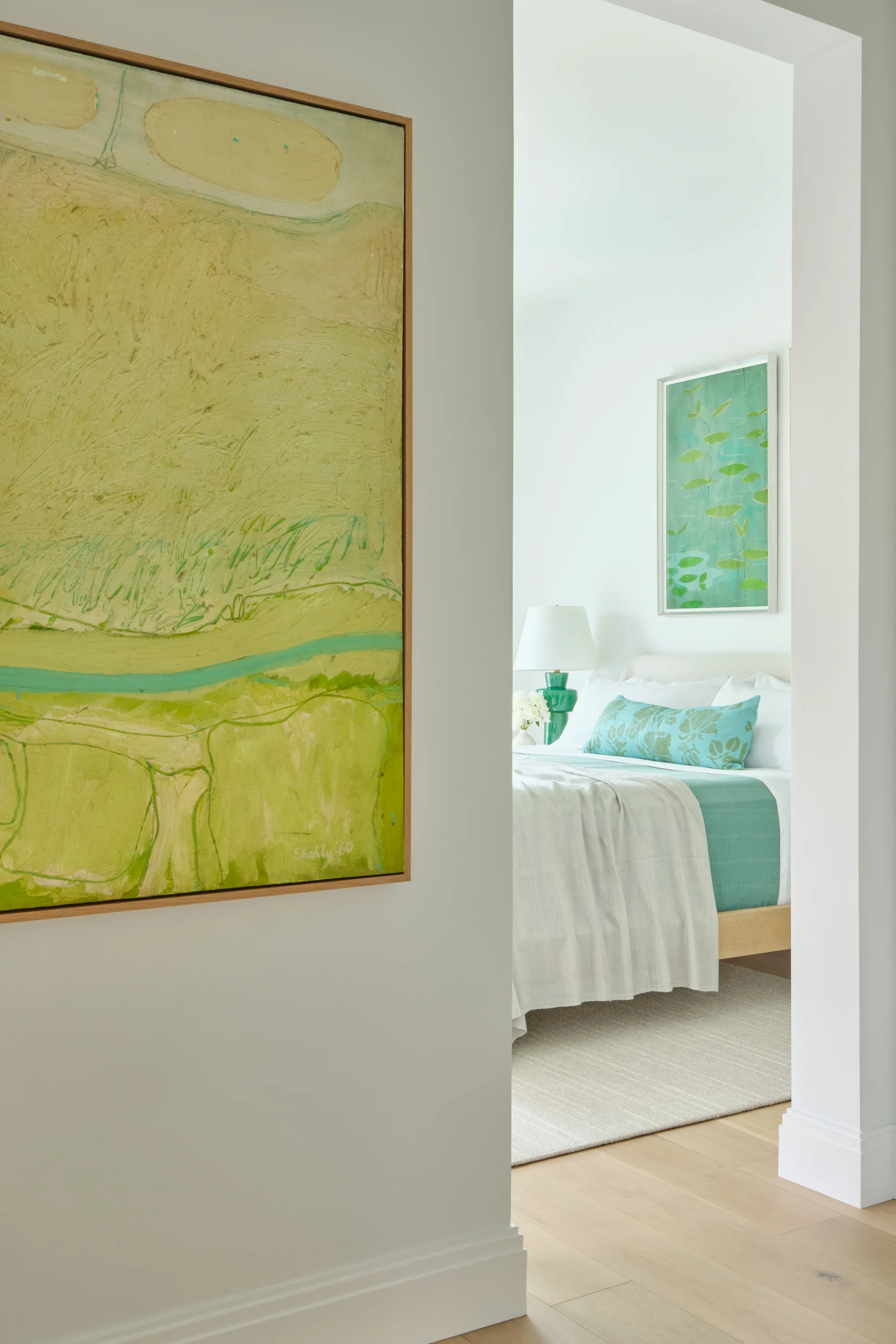 View through a doorway showing a bedroom with a white bed topped with green and blue pillows and a matching throw, a green lamp on the bedside table, light wood flooring, and two abstract paintings with green tones, one hanging on the bedroom wall and another partially visible on the adjacent wall.