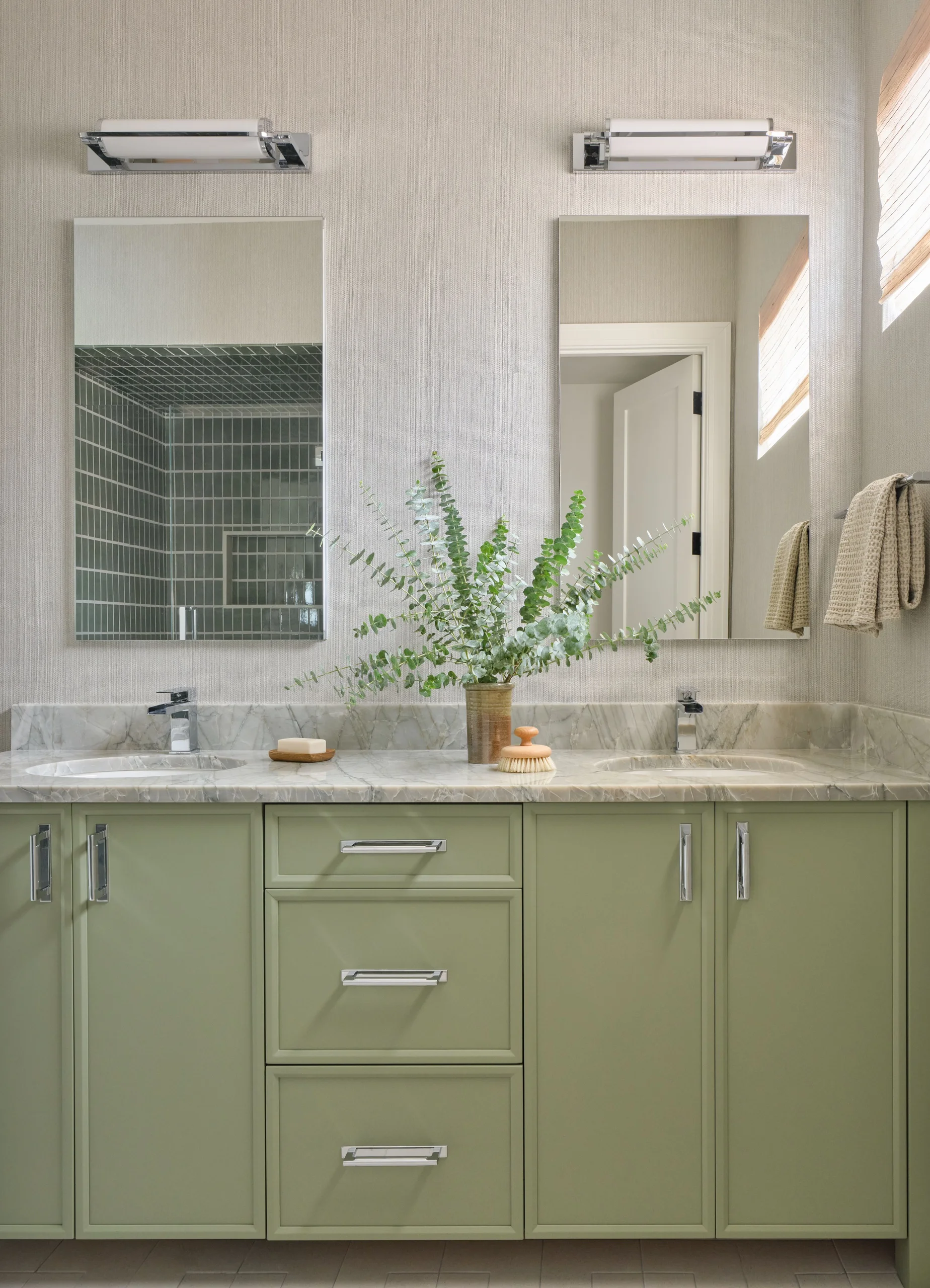 Bathroom vanity with light green cabinets, marble countertop, two under-mount sinks with chrome faucets, two rectangular mirrors, wall lights above them, and a vase with green leafy branches between the sinks.
