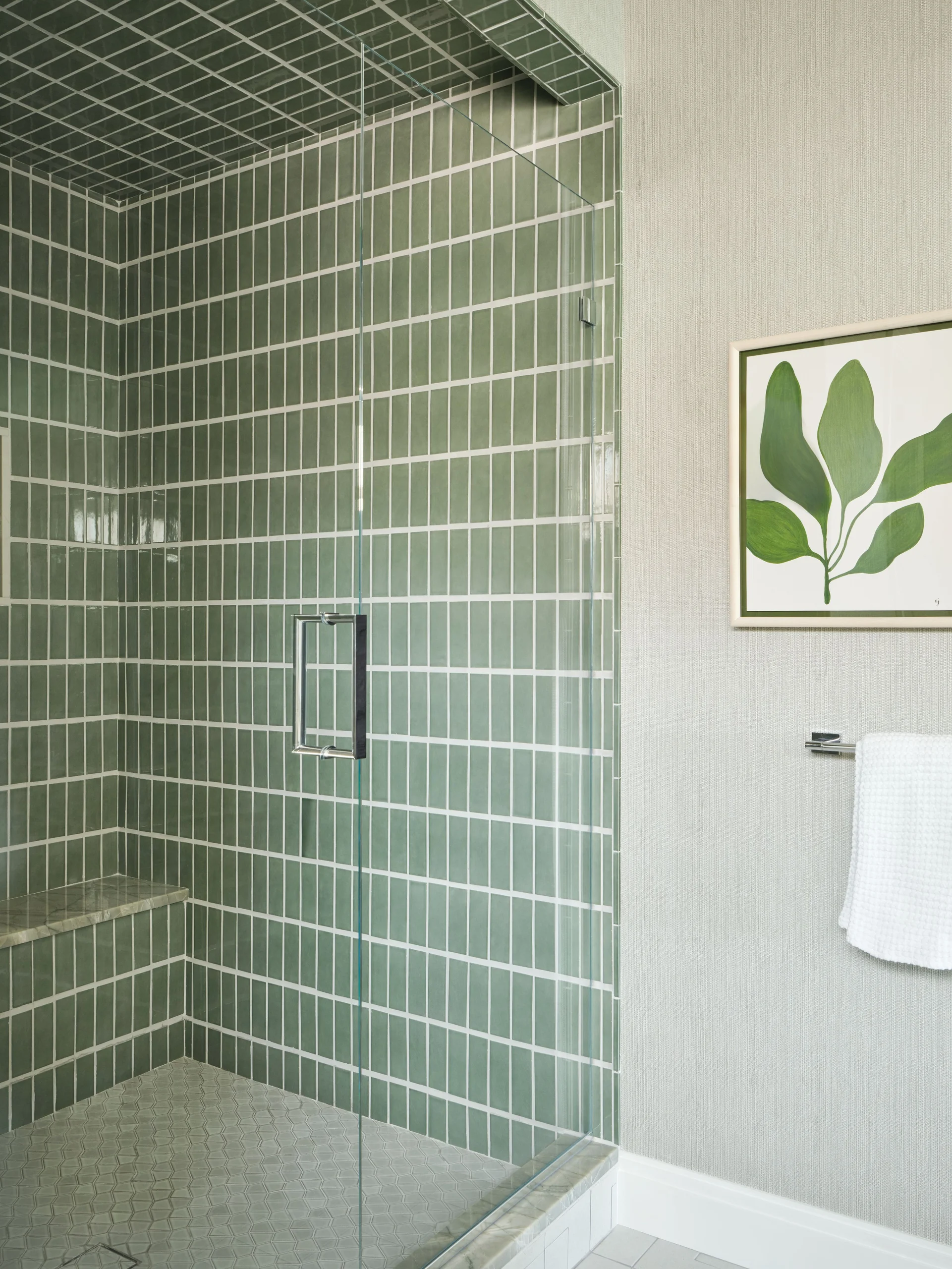 Modern shower area with vertical green tiles and white grout, featuring a glass door with metal handle and a built-in tiled bench, alongside a light gray textured wall with framed green leaf artwork and a white towel on a chrome towel bar.