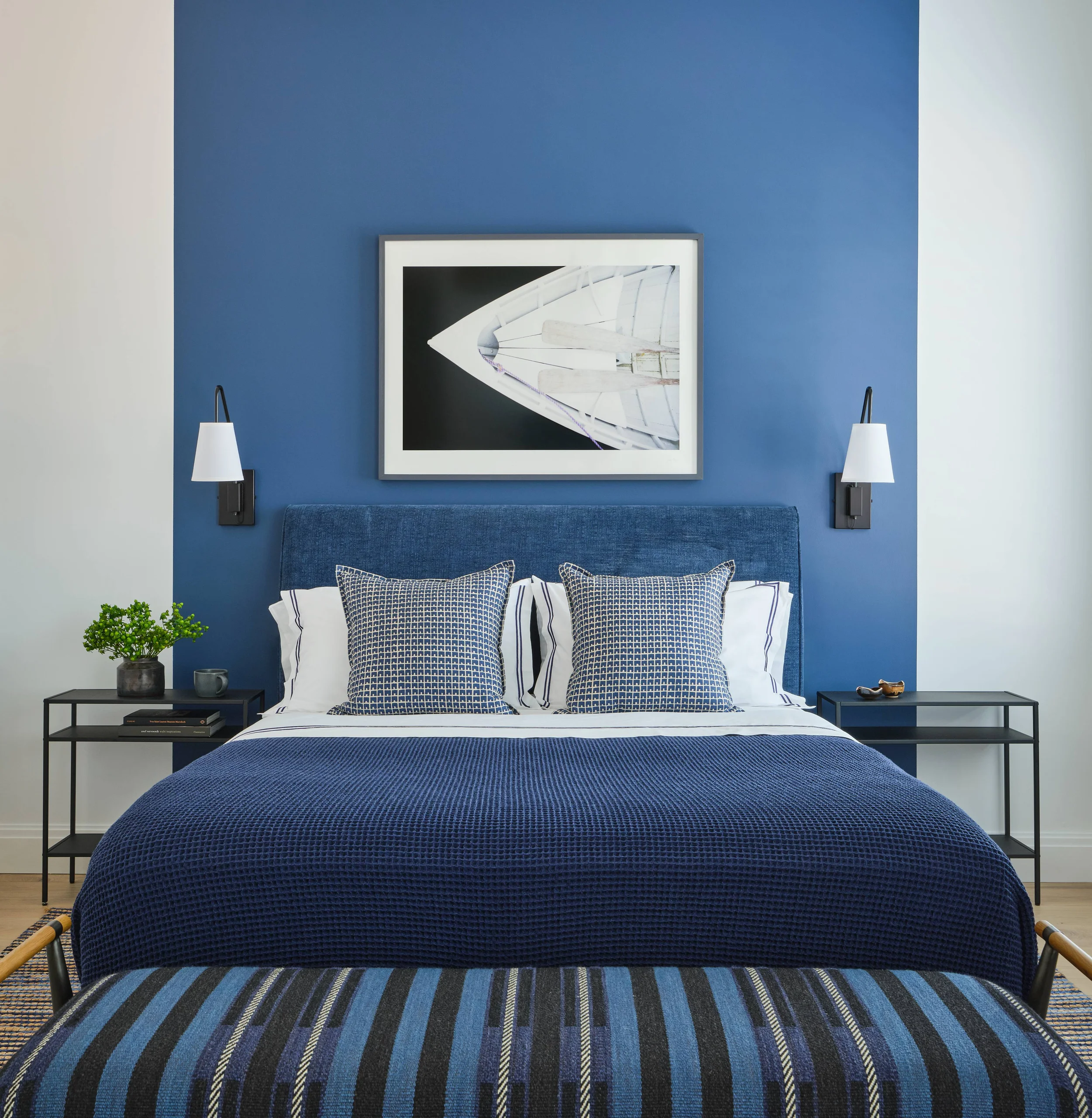 Bedroom with a navy blue accent wall behind a bed featuring a blue upholstered headboard, blue patterned pillows, white sheets with navy stripes, and a textured blue bedspread, flanked by black metal nightstands each with a wall-mounted white lampshade; a framed photo of a white boat is centered above the bed, and a blue striped bench is at the foot of the bed.