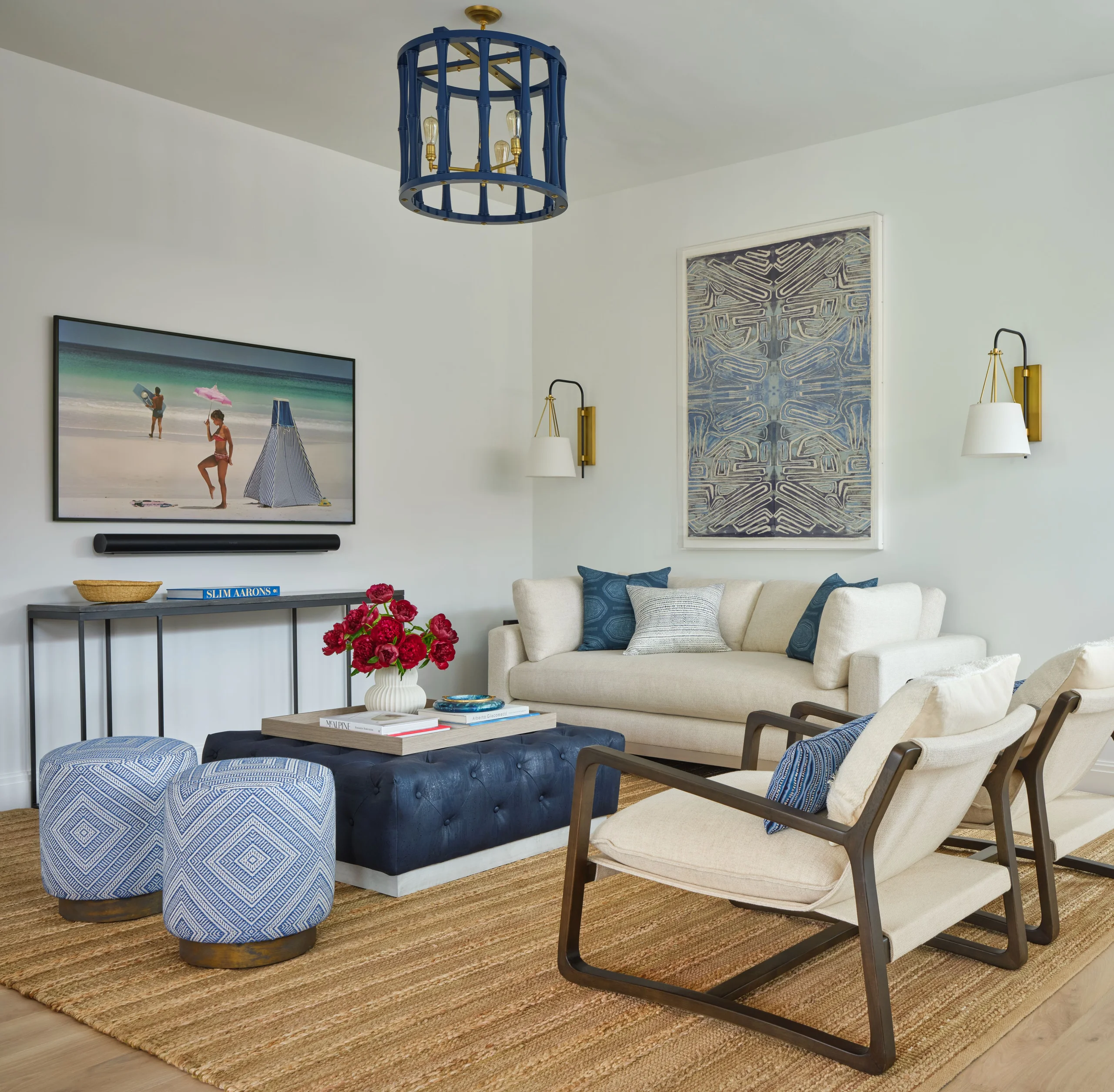 Modern living room with beige sofa adorned with blue and white pillows, two beige cushioned armchairs with dark wood frames, and two blue patterned ottomans. A navy tufted ottoman coffee table holds books and a vase with red flowers. A textured brown rug covers the wooden floor. Wall decor includes a framed abstract blue artwork and a beach scene photograph above a slim console table with books and a woven basket. Two wall sconces with white shades flank the artwork, and a blue ceiling light fixture hangs above.