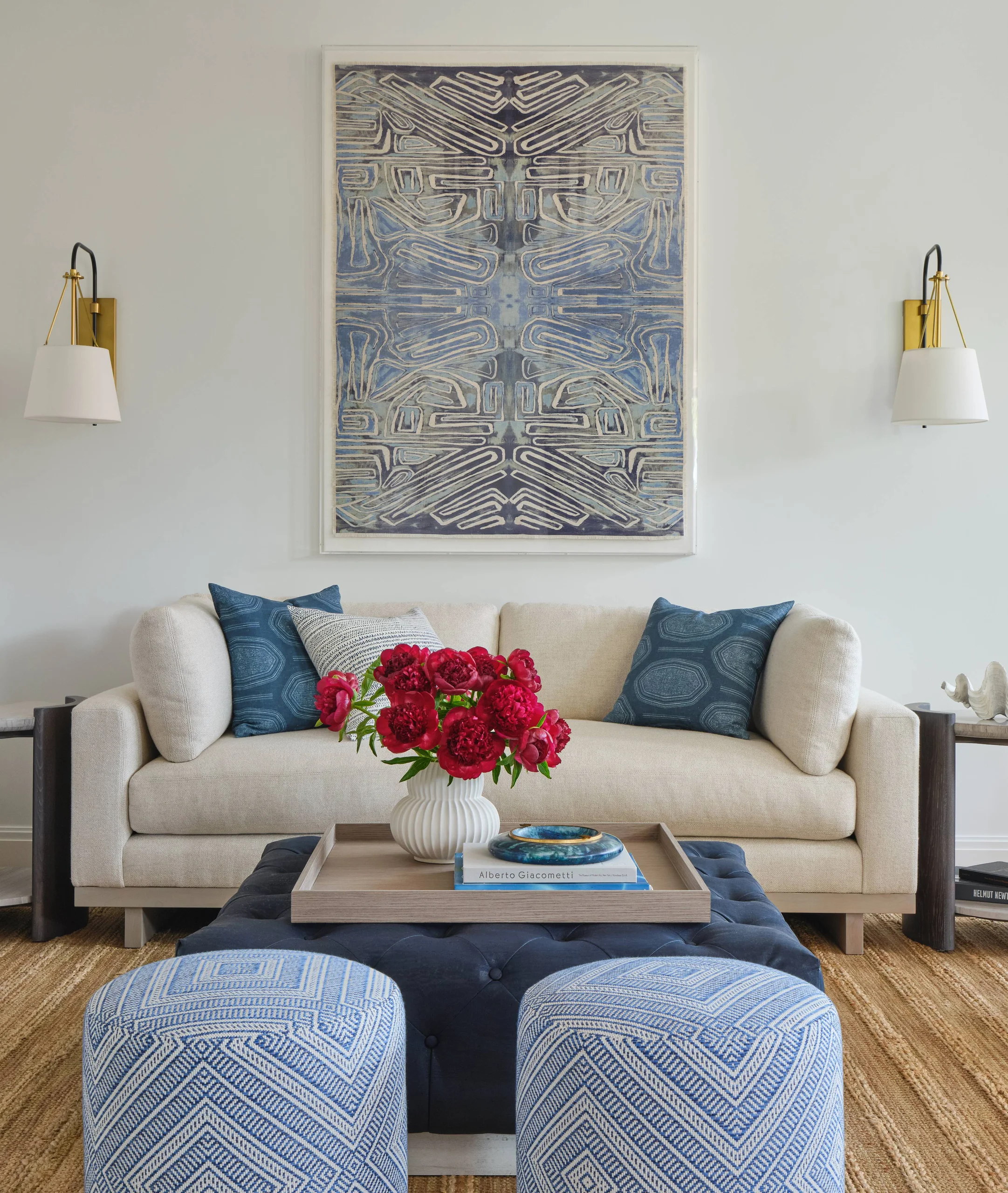 Modern living room with a beige sofa adorned with blue and white patterned cushions, a large abstract blue and white artwork on the wall, and two blue geometric-patterned ottomans in front on a natural fiber rug. A wooden tray on a dark tufted ottoman holds a white vase with red flowers and two books.