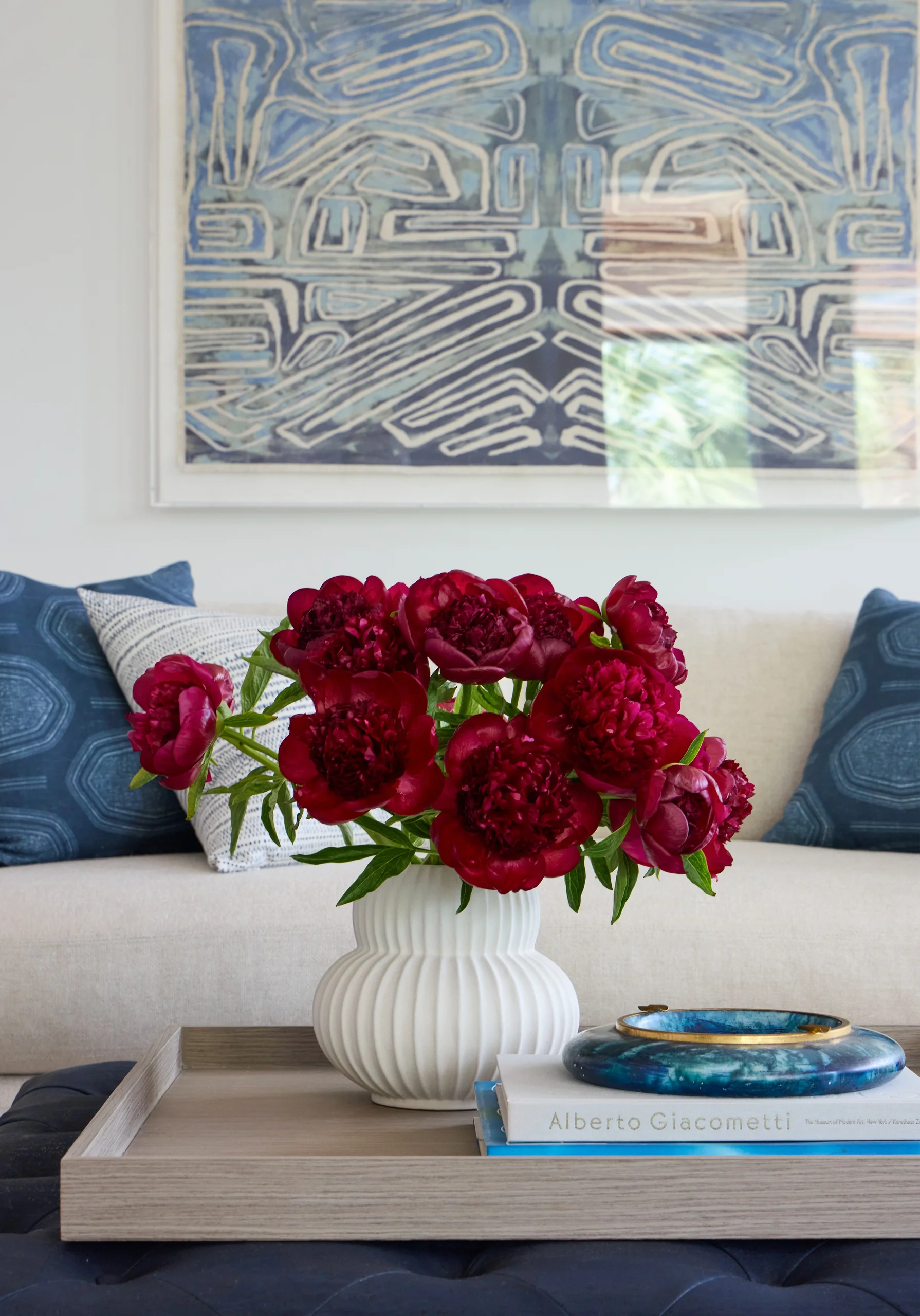 A white ribbed vase filled with vibrant red peonies sits on a wooden tray atop a blue tufted surface, with a book titled 'Alberto Giacometti' and a blue decorative object next to it, in front of a beige sofa with patterned blue and white pillows and an abstract blue artwork on the wall behind.