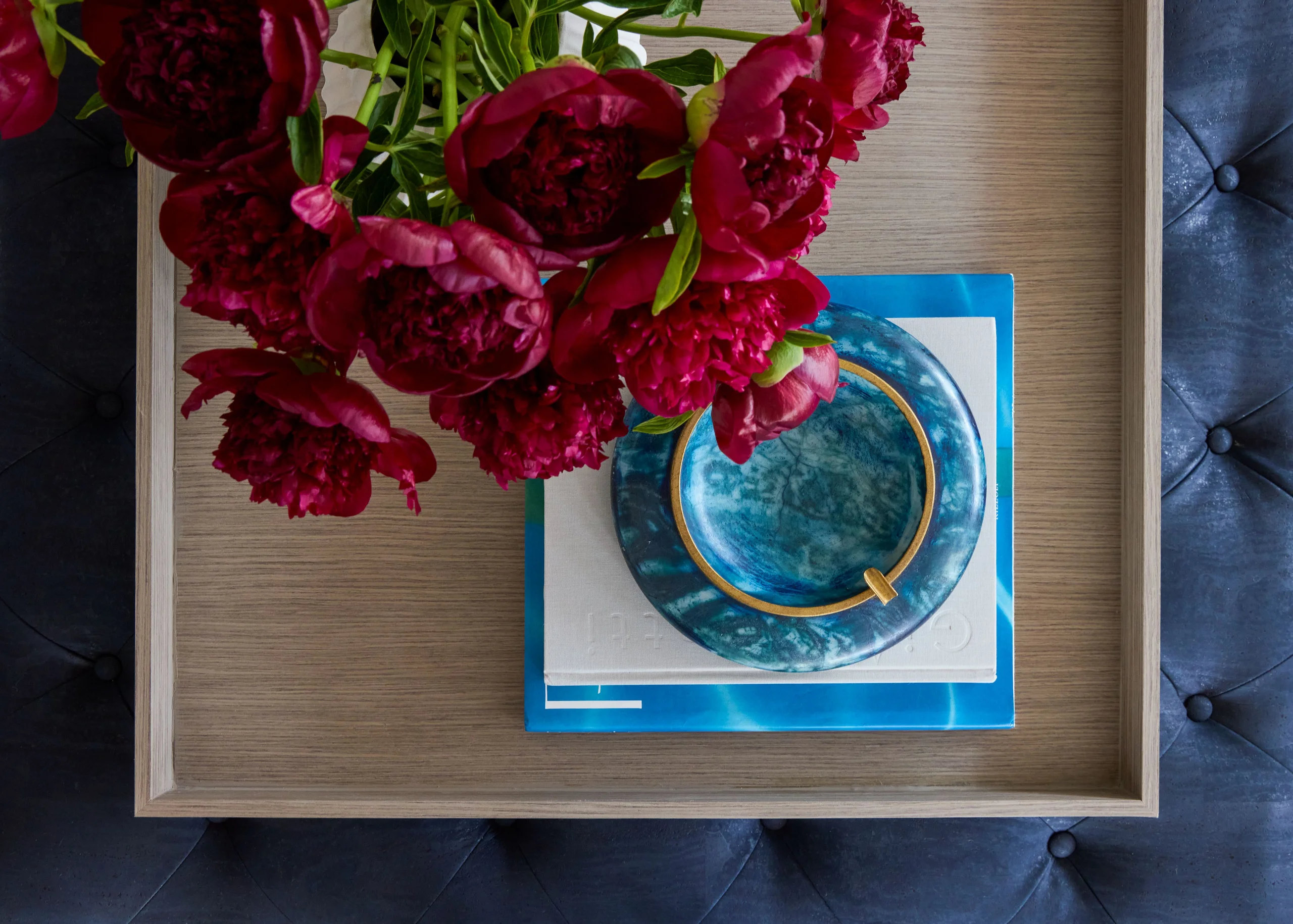 Top view of a wooden tray holding a stack of two closed books, one blue and one white, with a blue-green marbled ashtray resting on top, and a bouquet of deep red peonies partially covering the left side, all on a deep blue tufted surface.