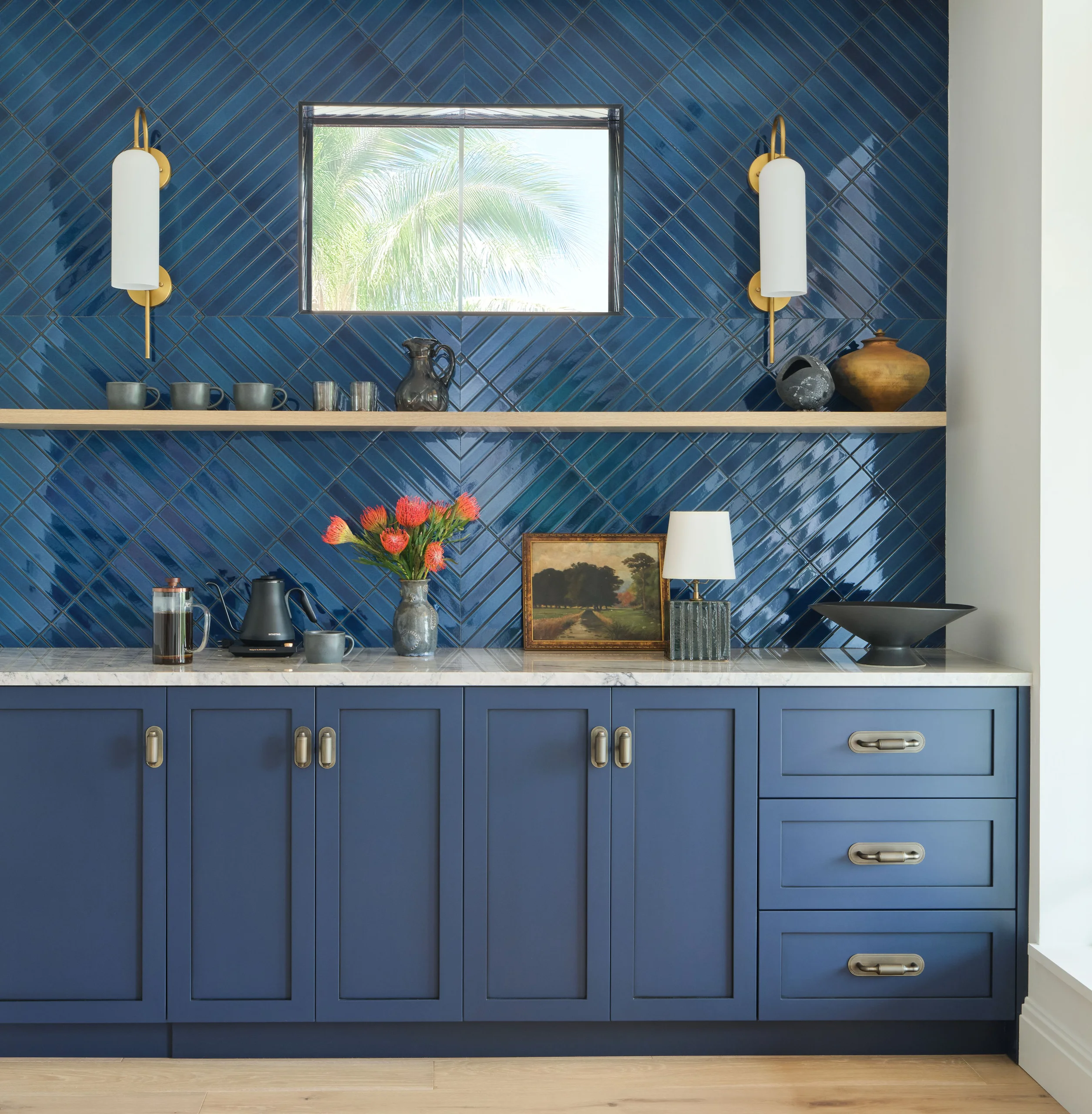 Kitchen area with blue cabinets and blue glossy herringbone tile backsplash, a small rectangular window showing palm tree outside, wooden shelf with cups and decorative items, marble countertop with coffee equipment, vase of orange flowers, framed landscape painting, table lamp, and black bowl, flanked by brass and white wall sconces.