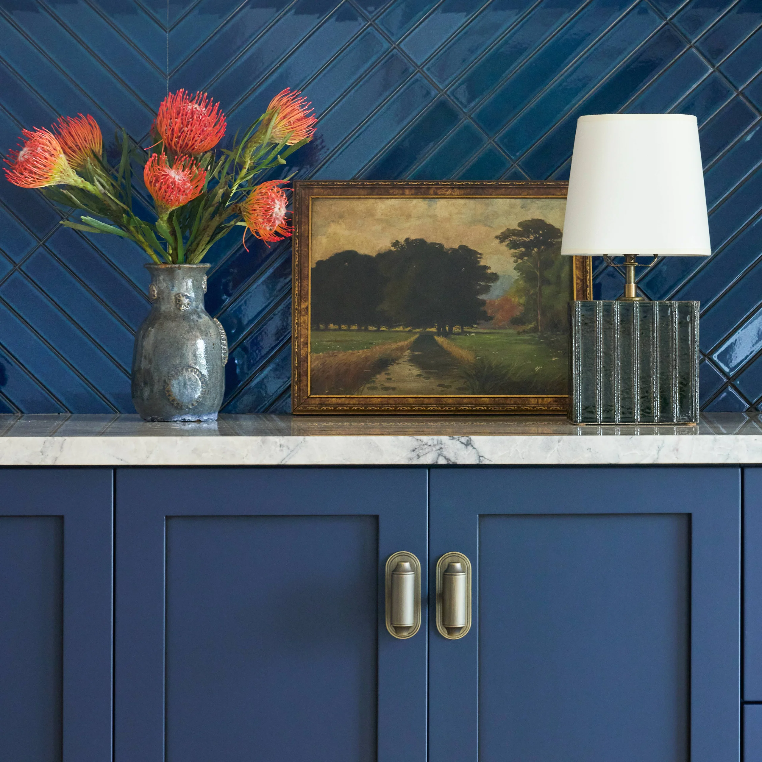 Blue cabinet with brass handles and a marble countertop holding a textured vase with red-orange flowers, a framed landscape painting of a tree-lined path, and a table lamp with a white shade and ribbed green base against a blue tiled wall.