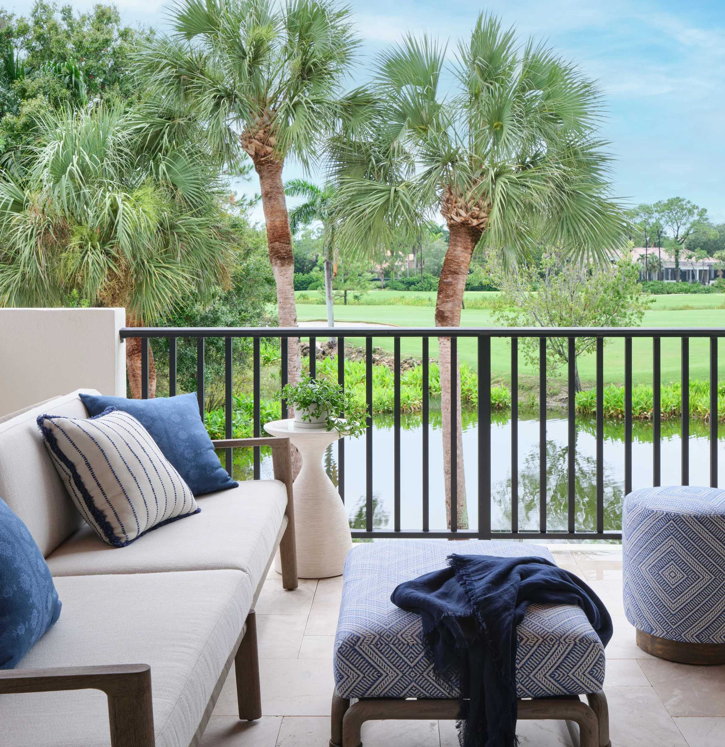 Outdoor balcony seating area with beige cushioned sofa and blue pillows, a small beige table with a potted plant, and a blue patterned ottoman with a dark blue cloth draped over it, overlooking a pond, palm trees, and green landscape under a partly cloudy sky.