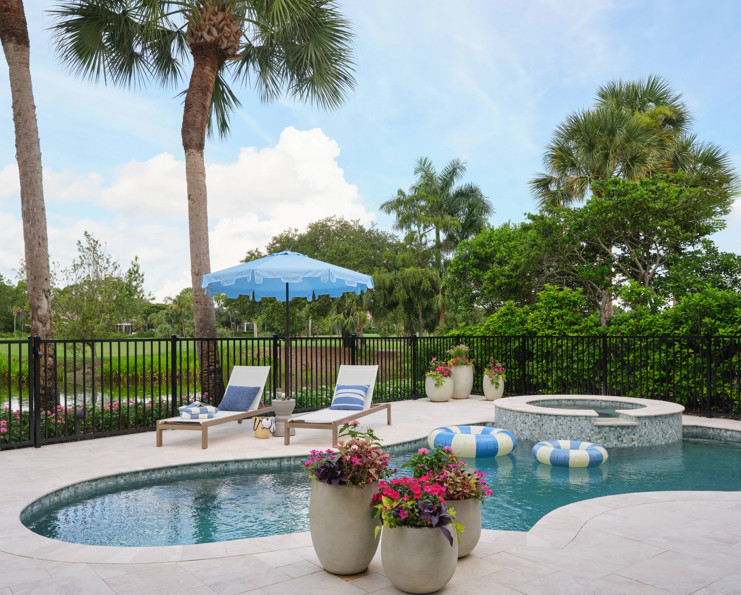 Outdoor pool area with three large pots of colorful flowers in the foreground, a kidney-shaped pool and round spa with mosaic tile surround, two lounge chairs with blue and white cushions under a blue umbrella, and palm trees and greenery beyond a black metal fence under a partly cloudy sky.