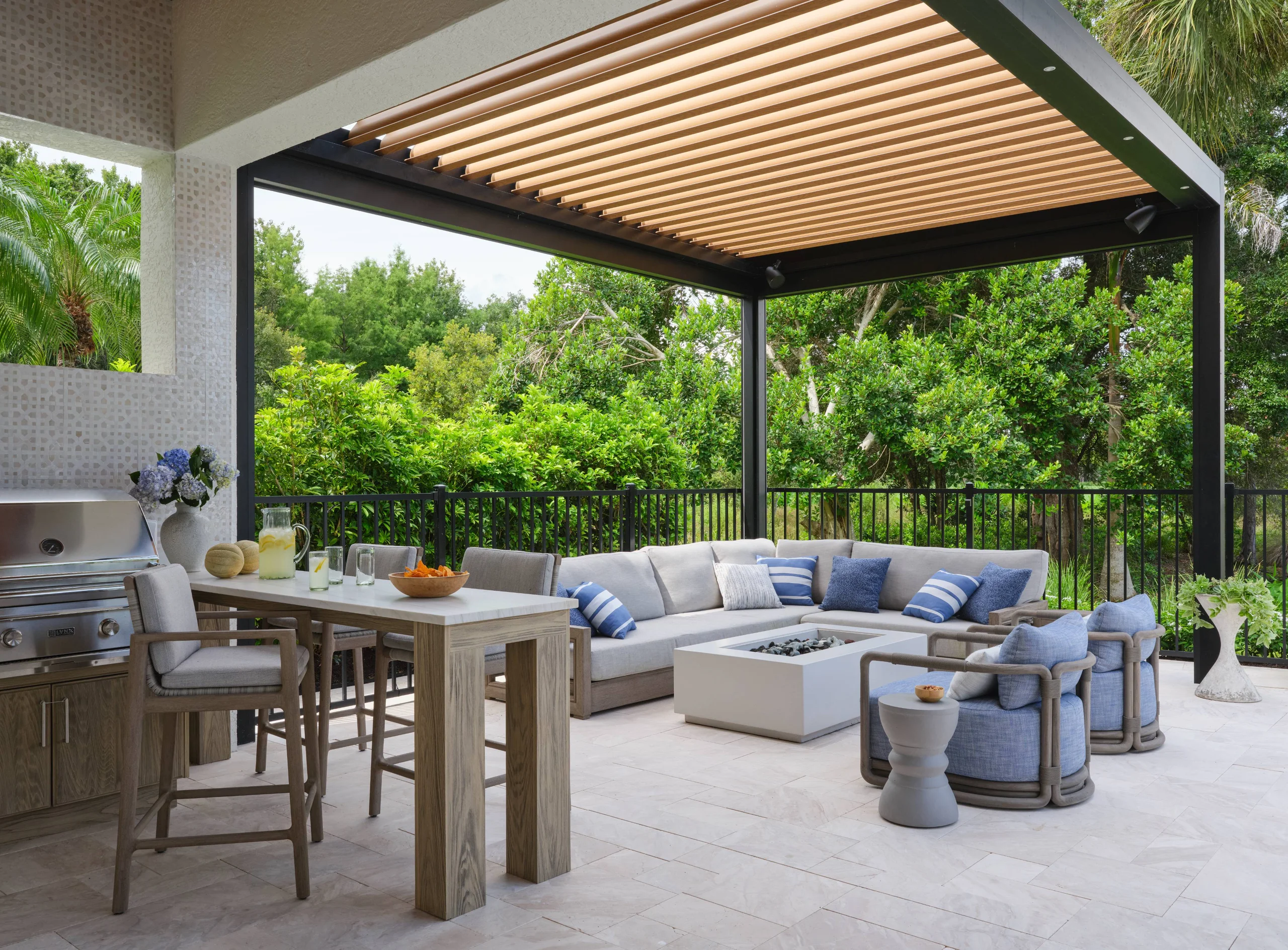 Modern outdoor patio with a beige sectional sofa adorned with blue and white striped and solid pillows, two light blue cushioned armchairs, and a white rectangular fire pit table under a pergola with wooden slats ceiling, surrounded by green trees and black metal fencing. A tall wooden table with two cushioned bar stools and a pitcher of lemonade, glasses, and a bowl of snacks is next to a built-in stainless steel grill.