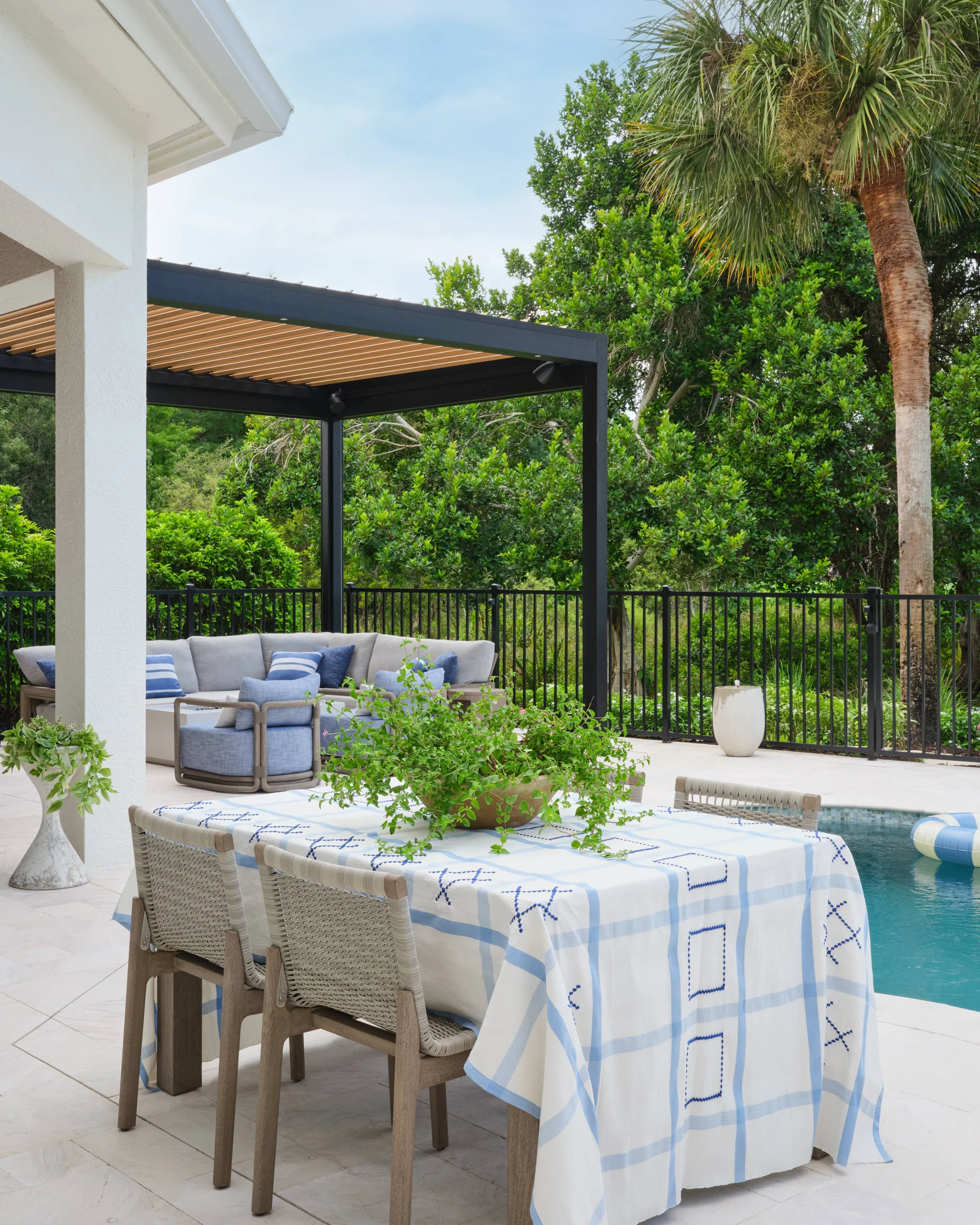 Outdoor patio area with a table covered by a white and light blue checkered tablecloth surrounded by woven-backed chairs, a potted green plant centerpiece on the table, a nearby swimming pool with a floating ring, a shaded seating area with gray cushions and blue pillows under a pergola, and lush green trees and a palm tree in the background.