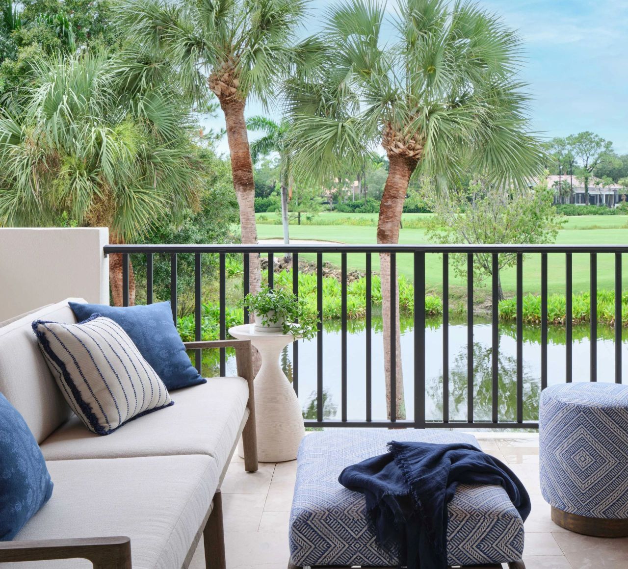Balcony with beige cushioned sofa with blue and white striped pillows, a small white side table with a green plant, and a blue patterned ottoman with a dark blue throw, overlooking palm trees, a pond, and a green lawn under a blue sky.