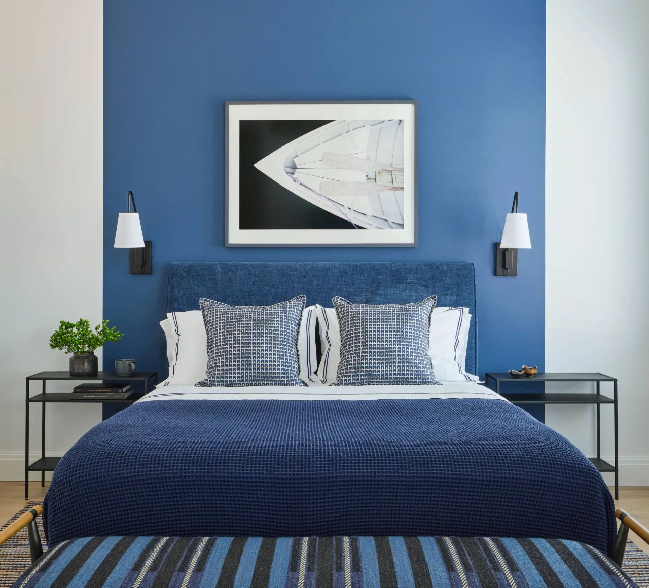 Modern bedroom with a blue upholstered headboard and a blue textured blanket on the bed, two patterned blue-grey pillows, white sheets with blue stripes, and a blue accent wall with a framed photo of a boat's bow above the bed; black wall lamps with white shades are on either side, with black metal side tables below, one holding a small plant and cup, and the other holding two small decorative objects.