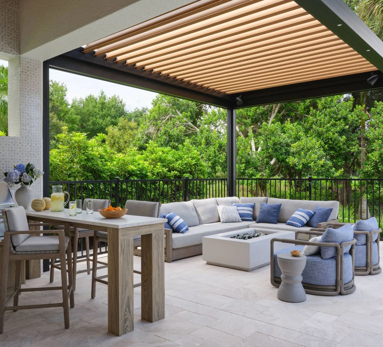 Covered outdoor patio with modern wooden pergola roof, light wood dining table with four chairs, a gray sectional sofa with blue and striped cushions around a white rectangular fire pit, two blue cushioned armchairs, a small round side table, and lush green trees in the background.