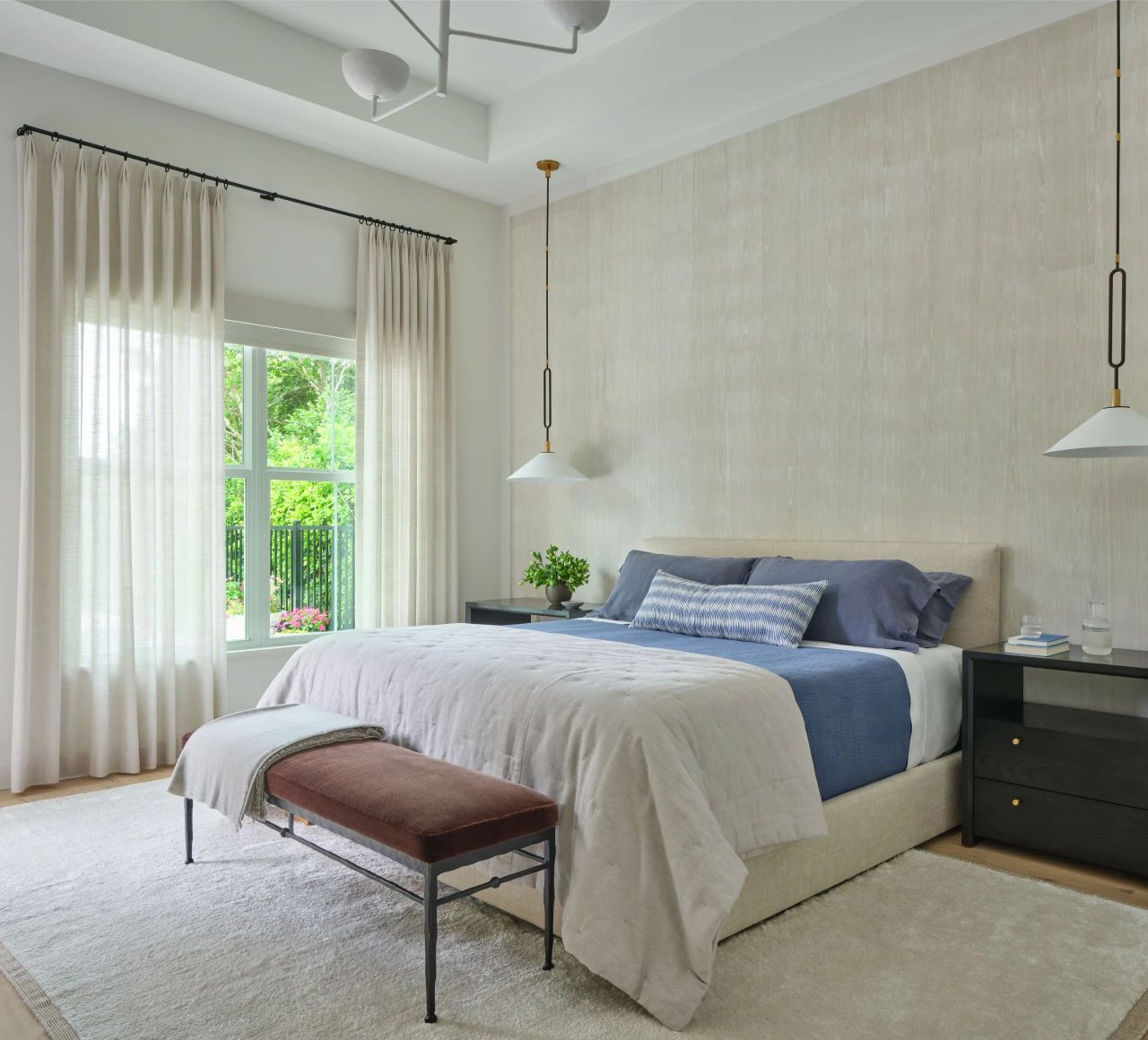 Modern bedroom with a large bed featuring beige bedding, blue blankets, and pillows, a brown bench at the foot, two dark nightstands with books and glassware, hanging pendant lamps, and a large window with sheer white curtains showing green foliage outside.